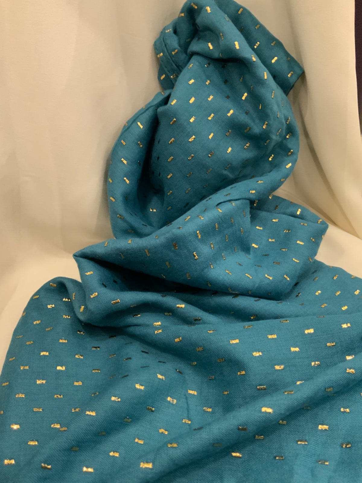 A teal blue fabric scarf or shawl draped over a cream-colored chair, decorated with a small repeating pattern of metallic gold embroidered figures.