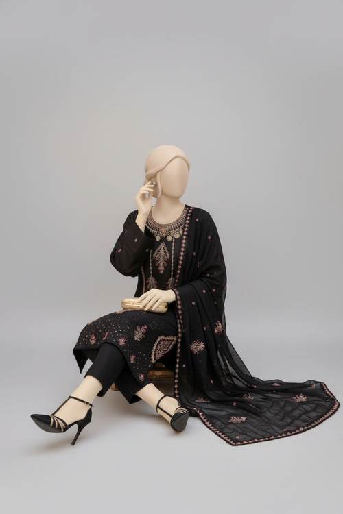A black three-piece traditional outfit displayed on mannequins in an ornate indoor setting with gold-framed furniture, red cushions, and decorative orange flower arrangements. One mannequin stands upright showing the full kurta, pants, and dupatta, while the other sits to show the draped fabric detail.