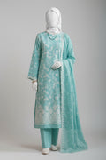 A woman wearing a mint green traditional outfit with white floral embroidery, a matching headscarf, and white gloves, standing in a styled studio setting with a terracotta wall, bamboo screen, and green leaves.