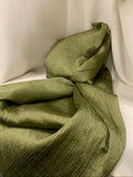 An olive green crinkled fabric, likely a lightweight scarf or wrap, draped and tied in a knot against a white fabric backdrop. A hand is visible at the top edge of the frame.