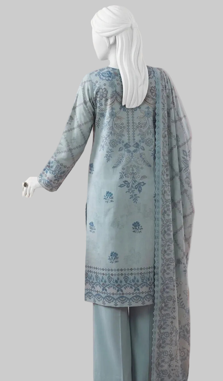 A woman wearing a light blue traditional outfit with intricate blue embroidery and a matching dupatta, shown from the back against a pink studio background with a wooden table and chair.