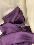 A close-up of draped purple fabric with a subtle crinkled texture, arranged against a soft off-white background.