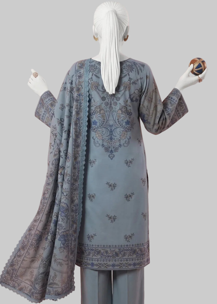 A woman wearing a three-piece traditional outfit stands with her back to the camera. The outfit consists of a dusty blue-gray kurta with intricate gold and brown floral embroidery, matching trousers, and a coordinating dupatta.